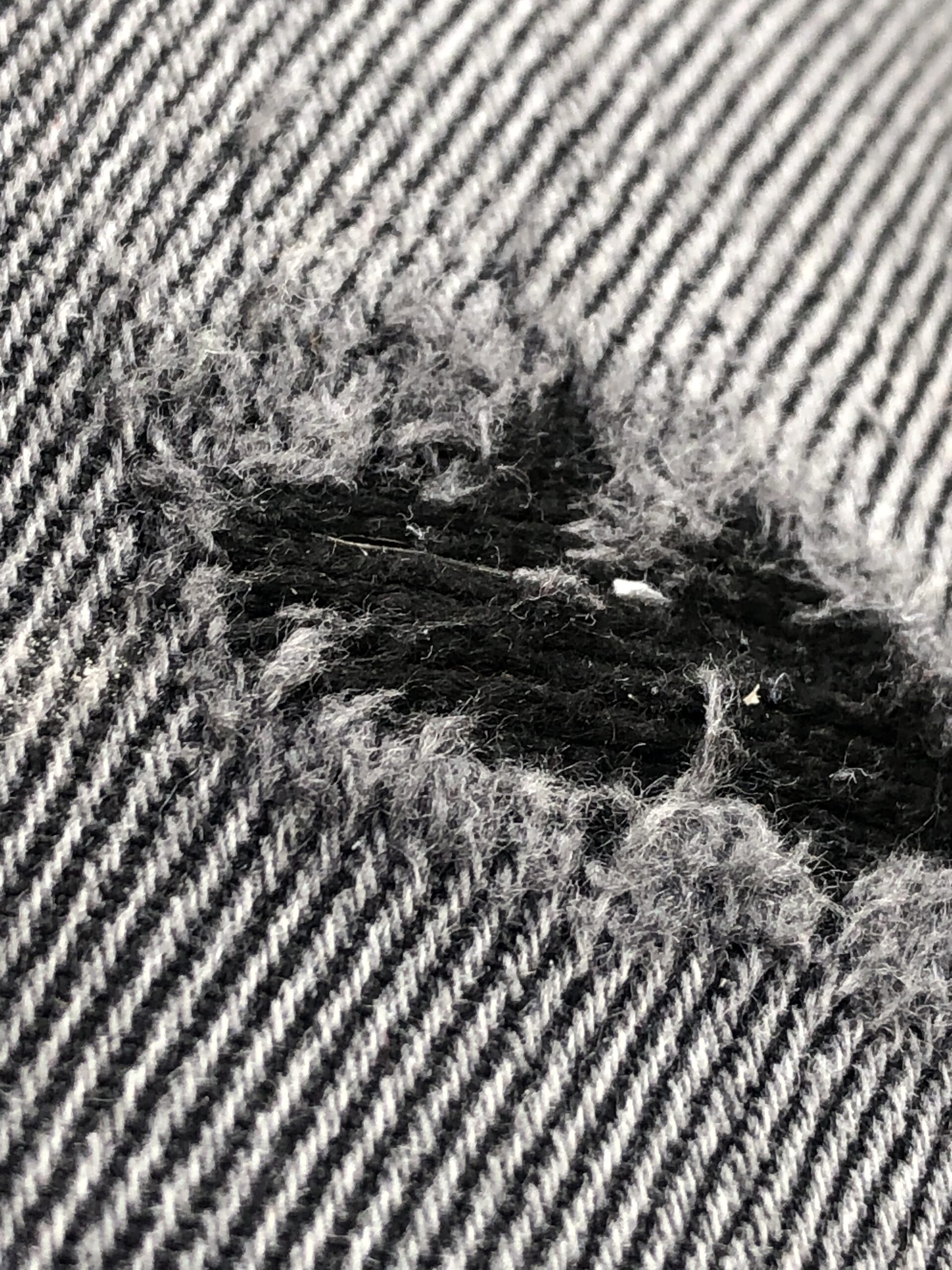 Loch in dem Textil einer schwarzen Jeanshose. Das regelmäßige Kettmuster wird unterbrochen von dem aufgerissenen Gewebe